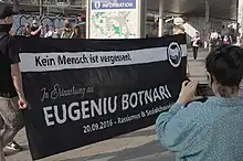 Zwei Personen halten ein schwarzes Transparent auf einem größeren Platz. Dahinter stehen und bewegen sich weitere Personen, davor fotografiert eine Person das Transparent auf dem steht „Kein Mensch ist vergessen! In Erinnerung an Eugeniu Botnari. 20.09.2016 – Rassismus & Sozialchauvinismus tötet!“