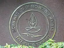 Ein Foto von einem messingfarbenen runden Emblem auf rotem Stein. Mittig eine stilisierte Feuerschale mit Rose darunter ein Blitz. Außen Kreisrunde Schrift Indian Institute of Technology unten indische Schriftzeichen.