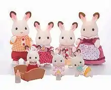 Auf dem Foto sind sieben Spielfiguren zu sehen, die eine Hasenfamilie darstellen. Die Eltern sind mehrere Zentimeter groß, die beiden Kinder etwas kleiner. Die Hasen haben ein weißes Fell, pinke Nasen, schwarze Knopfaugen und rosa Innenohren mit einer weiß-braunen Umrandung. Sie tragen von links nach rechts: Ein orange-weiß kariertes Hemd mit zwei roten Knöpfen sowie rote Shorts. Ein rotes Kleid mit weißen Punkten und einen orange-blumenförmigen Anstecker unterm Kragen. Ein weiß-rot kariert-gestreiftes Hemd mit zwei roten Knöpfen und beige Shorts. Ein lila Kleid mit einer dunkelroten Schleife unterm Kragen sowie eine weiße Schürze mit roten Punkten. Vor der Familie sind drei deutlich kleinere Baby-Hasen platziert. Sie tragen von links nach rechts ein rotes Kleid, einen rosa Strampler sowie eine orange Hose mit weißen Blumen am oberen Rand. Das erste Baby steht in einer Wiege, daneben befindet sich eine Babyflasche mit rosa Deckel. Das zweite Baby hat eine Flasche mit gelbem Deckel, das dritte einen hellblauen Schnuller im Mund.
