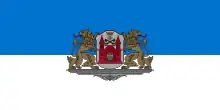Eine blau-weiße Flagge mit einem Wappen in der Mitte. Das Wappen hat eine rote Burg mit einem Löwenkopf in dessen Zentrum sowie eine Krone, ein Kreuz und zwei überkreuzte Schlüssel. Das Wappenschild wird von zwei braunen Löwen gehalten, die ihre Köpfe nach außen gedreht haben und ihre Zunge ausstrecken. Wappen und Löwen stehen auf einem grauen Podest.