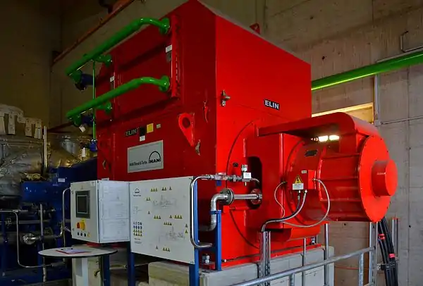 Synchrongenerator im ecki­gen rot lac­kier­ten Ge­häu­se mit bürs­ten­lo­ser Er­re­ger­ein­rich­tung; Er­re­ger­ma­schi­ne mit Per­ma­nent­mag­net­er­re­gung rechts am Ge­häu­se an­ge­flanscht; in den Edelstahl­lei­tun­gen die Ölschmierung; in den grü­nen Lei­tun­gen die Glykolkühlung mit in­ter­ner Luft­zirkulation