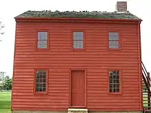 Zweistöckiges Haus mit roter Holzverschalung. Das Gebäude hat drei Sichtachsen und einen Kamin. Es ist schlicht und weist keine Schmuckelemente auf.