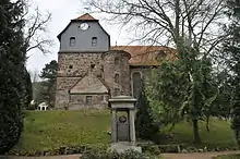 Blick aus Dorfrichtung mit vorgelagertem Kriegerdenkmal, in der Mitte des Kirchengebäudes der Treppenturm