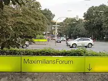 Straßenszene, im Vordergrund eine Reihe gelber Leuchtbausteine mit schwarzer Aufschrift "MaximilansForum" und Pfeil nach unten