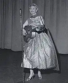 Eine Frau mit kurzen blonden Haaren in Tüllkleid und Stöckelschuhen, mit einem Schal um den Hals steht vor einem Mikrofon auf einer Bühne; sie spielt Gitarre und singt.