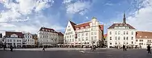 Frontale Farbfotografie von zwei hellen Häuserfassaden, die diagonal verlaufen und sich in der Mitte treffen. Einige Außenterrassen mit Menschen sind vor den Häusern und ein paar Menschen laufen über den Platz im Vordergrund. Hinter dem rechten Haus ist eine Turmlaterne zu sehen.