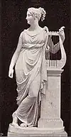 Statue von Terpsichore, Frontalansicht; sie lehnt sich an ein Podest links von ihr, auf dem eine große Lyra steht, diese hält sie zugleich mit der linken Hand