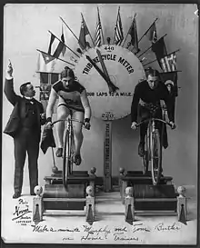 Ein Schwarzweißbild zeigt die Radsportler Charles Murphy und Tom Butler auf der freien Rolle. Im Hintergrund sieht man eine Anzeige, die die jeweils gefahrene Strecke anzeigt.
