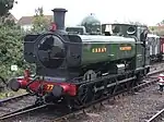A pannier tank locomotive is passing a station platform, running round its train which includes rolling stock visible in the background. The pannier tanker is green, apart from the black chimney, brass safety valve cover, red buffer beam, and grey coupling rods. Great Western is written on the side of the tank, and 7752 on a number plate on the cab
