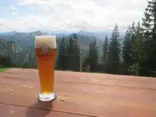 Im Vordergrund ein Weißbier im Glas auf einer Biertischgarnitur, im Hintergrund Bergpanorama